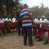 A group of Ugandan students sitting together  outside a classroom, receiving guidance and counseling from a teacher. The students are engaged, listening attentively and smiling as they learn life skills and personal development. This image represents TNYME Uganda’s mission to support disadvantaged children through mentorship, education and holistic development. Volunteers can help set up and manage a GoFundMe campaign to raise funds for building a permanent community school on 2 acres of land, directly impacting these children’s future.