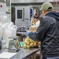 a man holds a bag of apples, women in the background prepares food containers