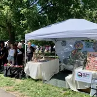 7 people standing under a tree, next to a white canopy, 10 foot square - Fabric dolls from Haiti are in a black wagon at left, metal art hangs in the canopy, tables are filled with bracelets, earrings, and other Haitian art products. It's a sunny day, with blue sky above the tree.
