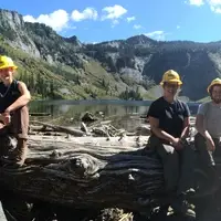 Two groups of three people sit side by side on a large log near an alpine lake; five are wearing yellow hard hats and one is wearing a white hard hat.
