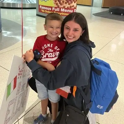 Young volunteer with short hair hugs small child at airport after volunteer opportunity in Italy.