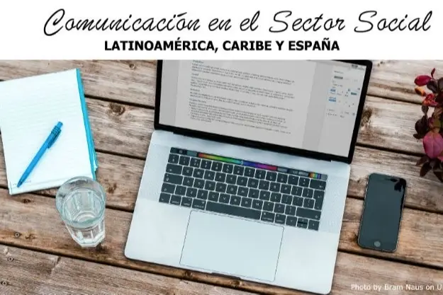 Una laptop en una mesa de trabajo junto a un cuaderno, un celular y un vaso de agua