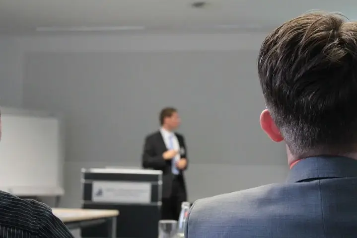 A person in a business suit stands at the front of the room and is watched by an audience.