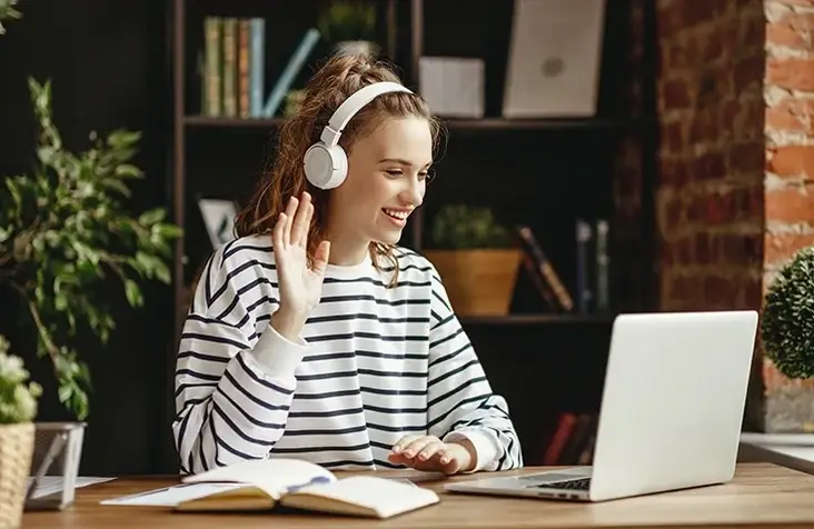 Young white girl sits at desk at home and waves to her laptop's camera during virtual volunteering session.