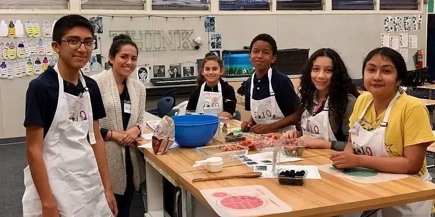 Children in aprons gather around a table with cooking ingredients. They are facing the camera.