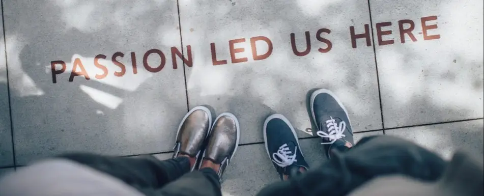 Two people's feet on a sidewalk with 'Passion Led Us Here' written on it.