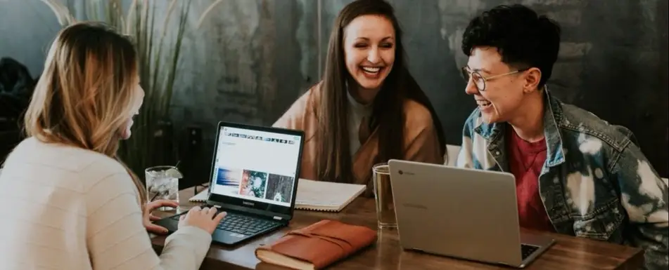 Three people sitting and laughing. They have their laptops and a notebook.