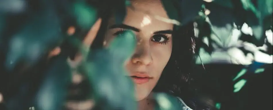 Woman with dark hair looks out from behind tree leaves.