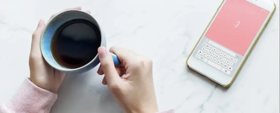 Hands holding a cup of coffee with a smart phone next to them.