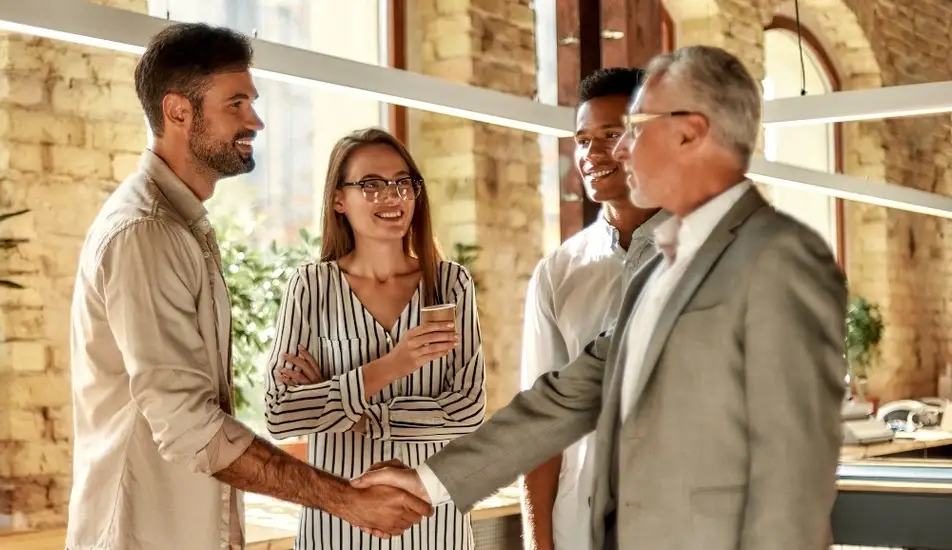 Dos personas dándose la mano y sonriendo mientras están de pie con sus colegas.