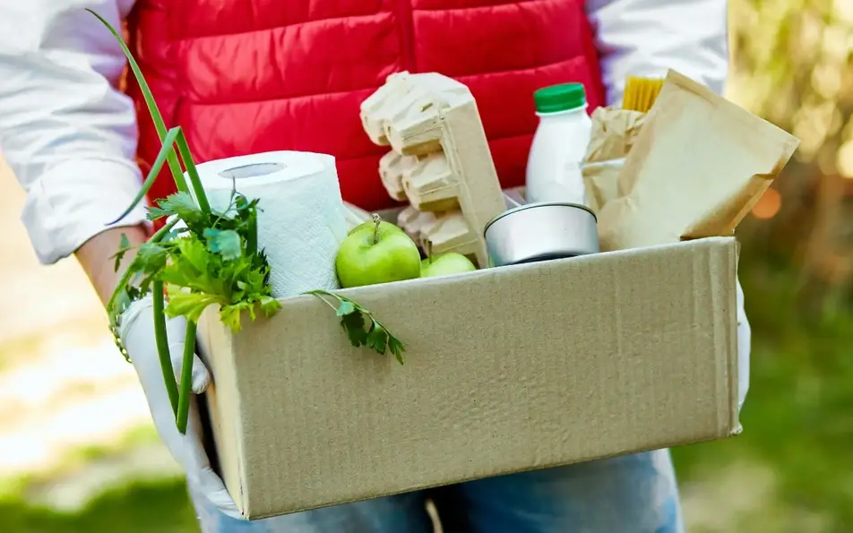 Volunteer brings groceries to someone in need, carrying a cardboard box of food and cleaning supplies.
