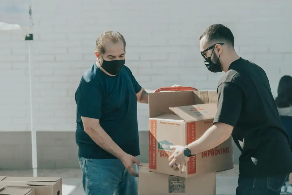 Imagem mostra 2 homens com mascara de proteção juntos ajudando no carregamento de caixas de papelão.