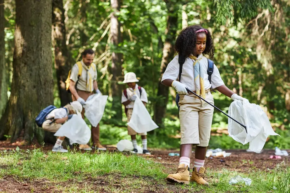 Young Black girl dressed in scouting uniform cleans up trash in the woods as part of community service with other den members.