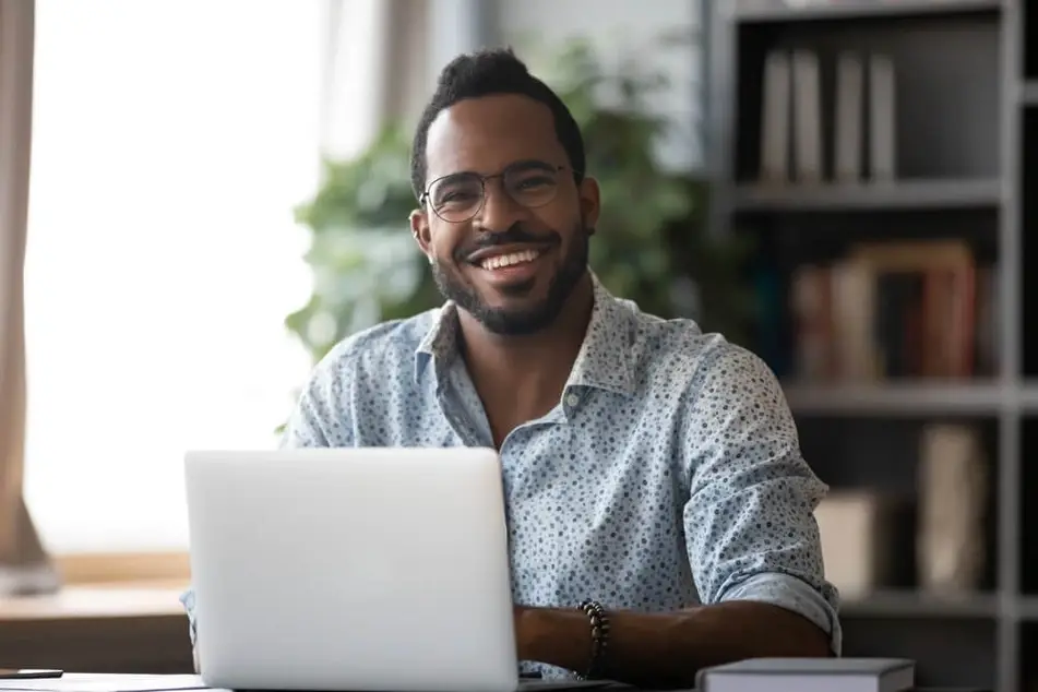 Black man smiles at a camera while working on his laptop at a desk