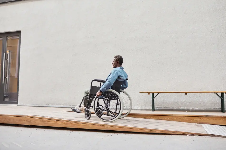 A Black man in a wheelchair uses a wooden ramp to enter a building.