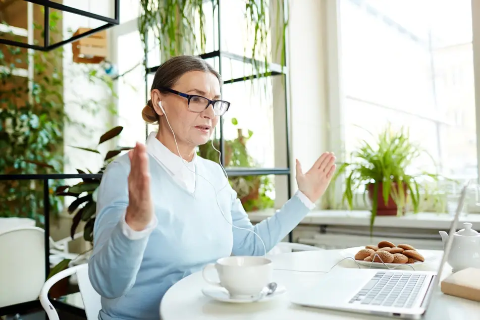 An older white woman wearing a blue sweater sits in a cafe as she takes a work meeting from her laptop. There are a tea cup and plate of cookies in front of her, with many green plants in the background.