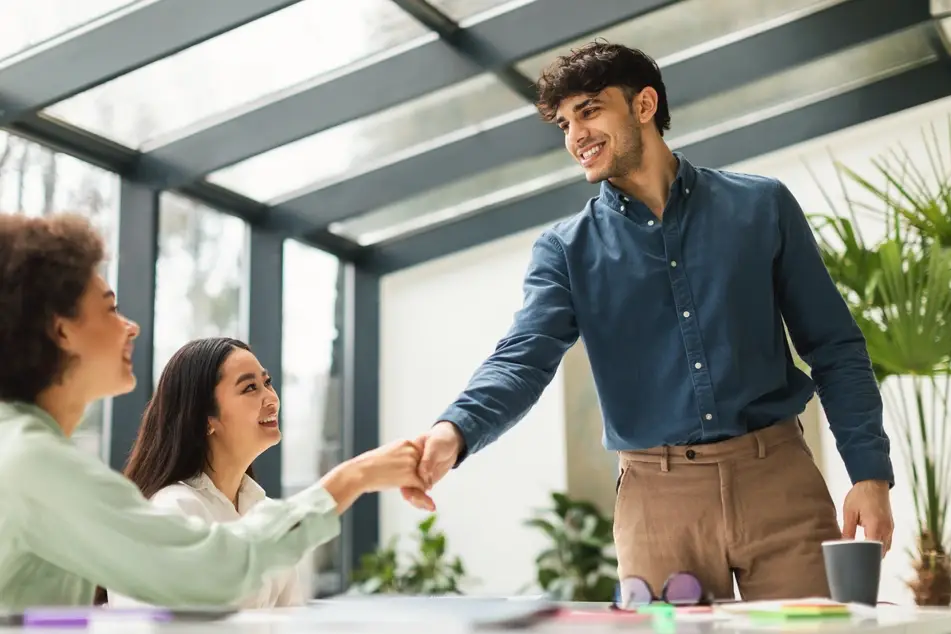 A young man with brown hair is standing over a long desk as he prepares for his performance review. He is shaking hands with a woman wearing a green blouse, while another woman with long brown hair smiles from the side. There are green plants in the background of a bright office space.