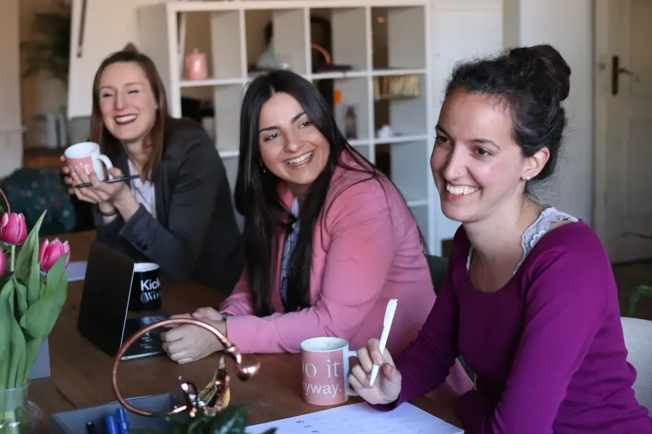 three women smiling at table with laptop and writing pad