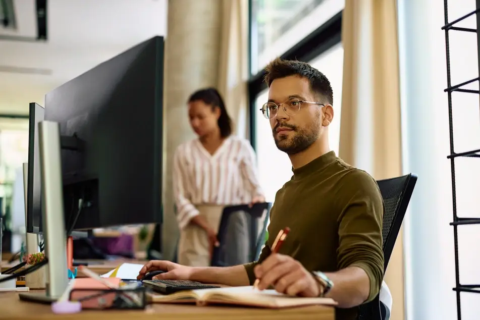 Young man writing notes while working on a computer in the office.