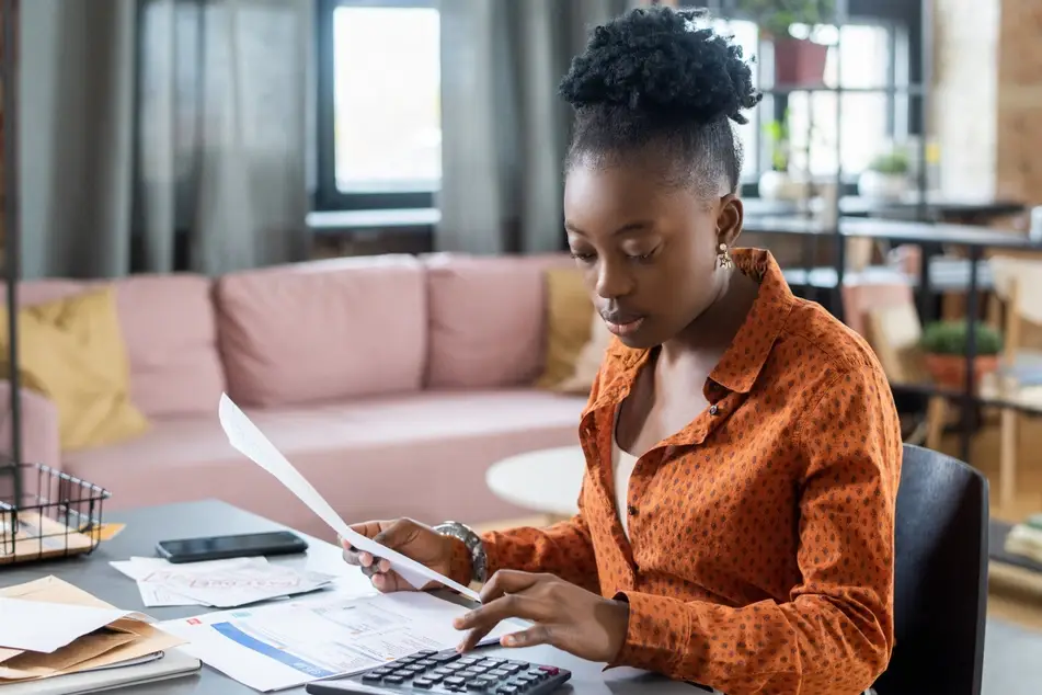 A Black woman sits at a desk in her living room crunching numbers as she considers when to ask for a raise. She is wearing an orange shirt, and there is a light pink couch with yellow pillows in the background.