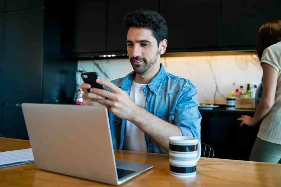 Hombre trabajando en una computadora mientras envía mensajes por celular