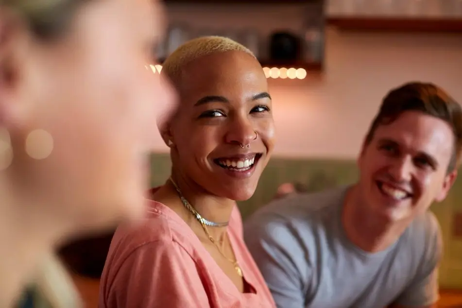 3 pessoas conversando e sorrindo. Um rapaz jovem ao fundo de uns 25 anos com uma camiseta branca, uma moça afrodescendente com cabelo raspado, com um sorriso muito livre, uma blusa rosa, colar e brinco com uns 30 anos, uma última moça jovem, também, com cabelo curto, camiseta. Todos com uma vestimenta casual e sorrindo.