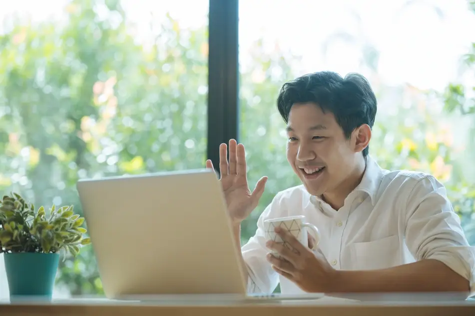young man waves at camera on laptop while smiling