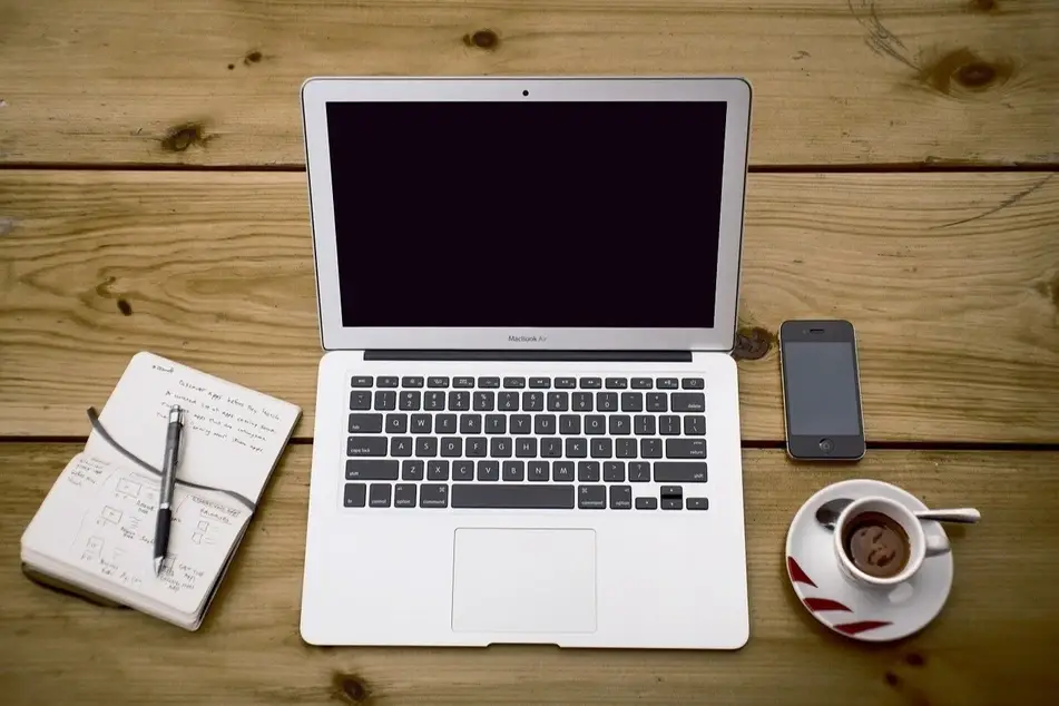 A laptop with a phone, cup of coffee, and notebook next to it.