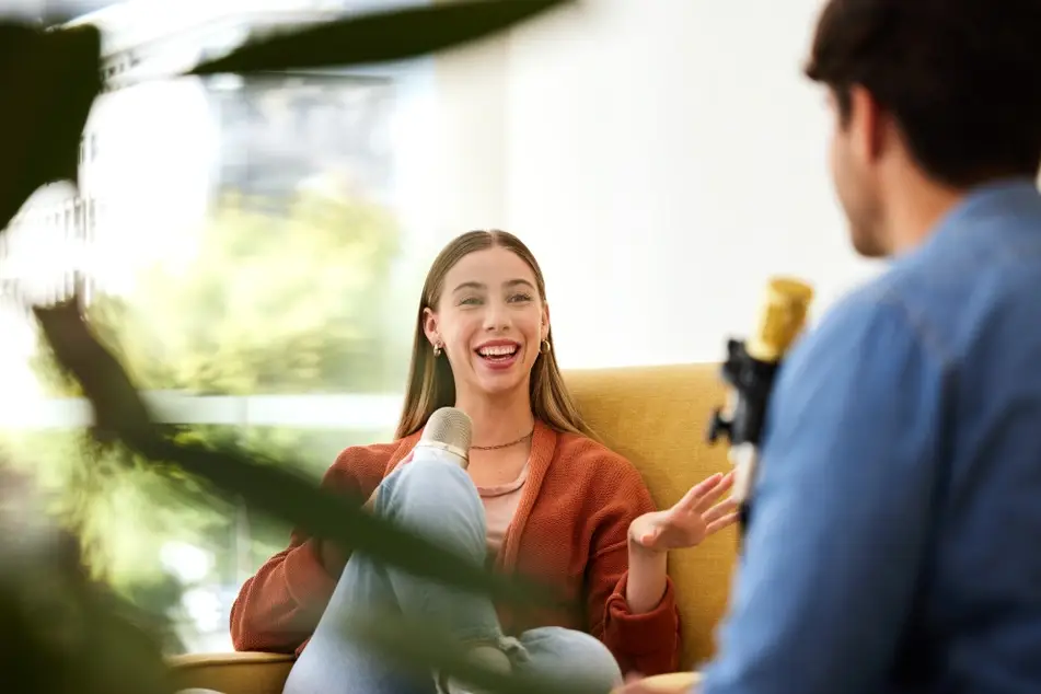 White woman holding a microphone smiles while speaking for a podcast interview while they both sit on a yellow couch.