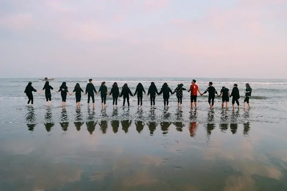 A group of 17 people holding hands in a long line at the beach