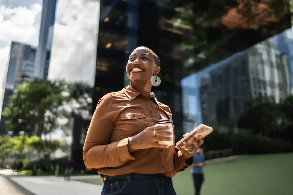 Black woman smiles up at sun, holding a coffee and her phone while walking outside.