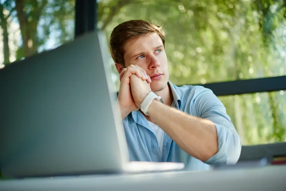 White man sits at office desk thinking, green trees in the background.