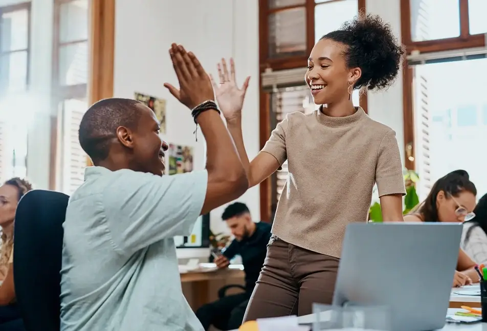 Two co-workers high-five at the office while other colleagues work around them