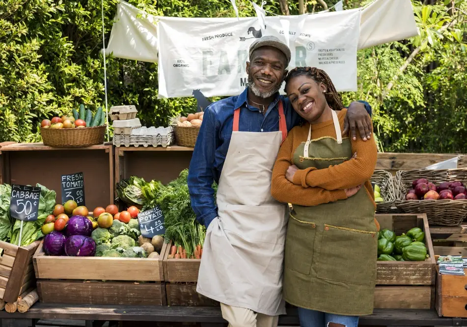 A Black man and woman stand together smiling in front of a fresh food stand to highlight the benefits of volunteering at a farm stand.