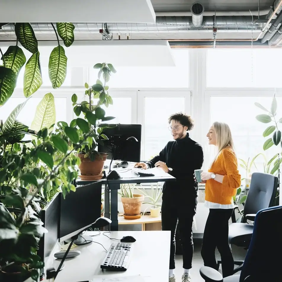 Two smiling co-workers look at a computer screen while surrounded by greenery.